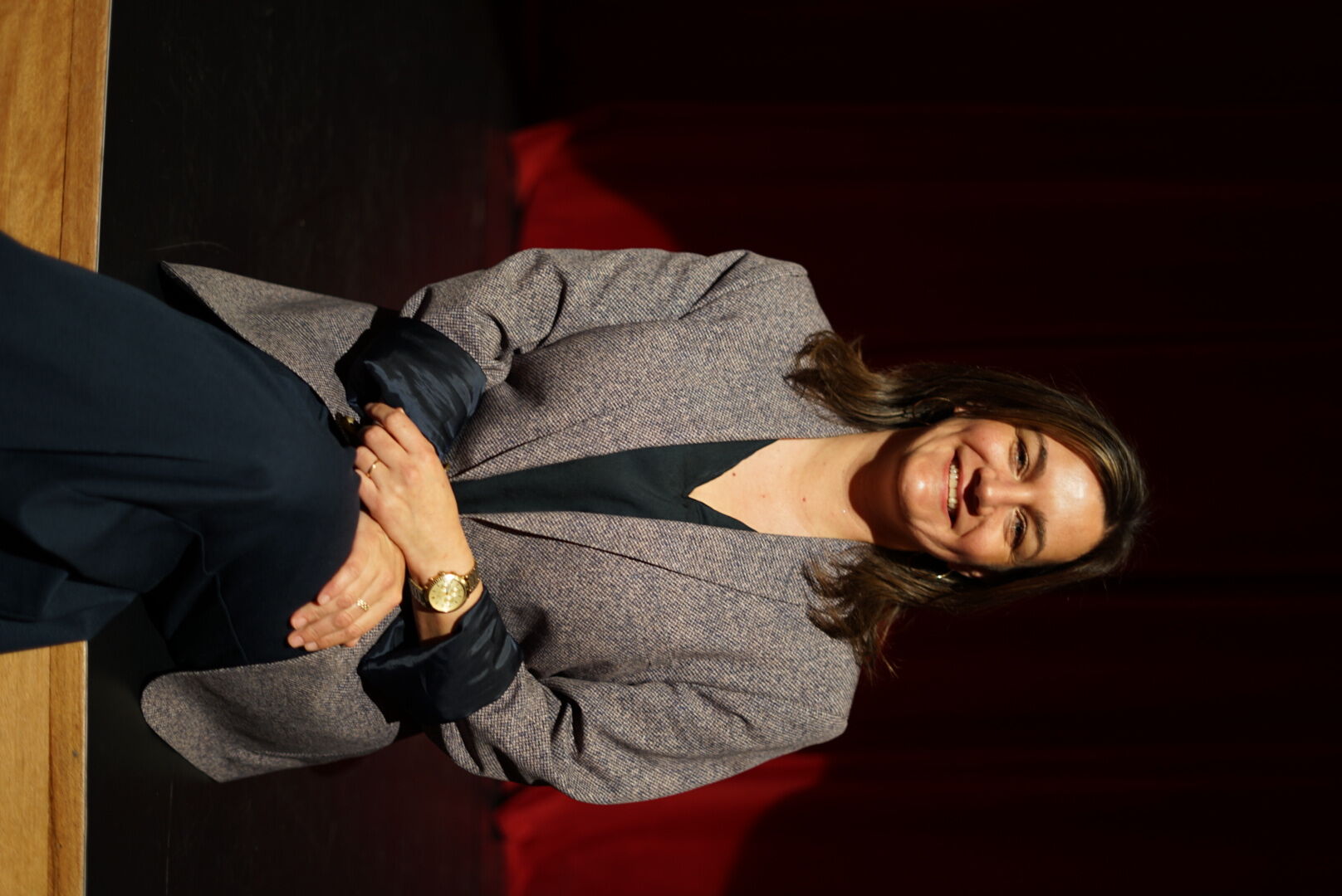 Professionele vrouw in een blazer, zittend op een podium met een rode achtergrond, glimlachend naar de camera.
