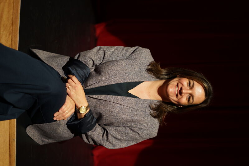 Professionele vrouw in een blazer, zittend op een podium met een rode achtergrond, glimlachend naar de camera.