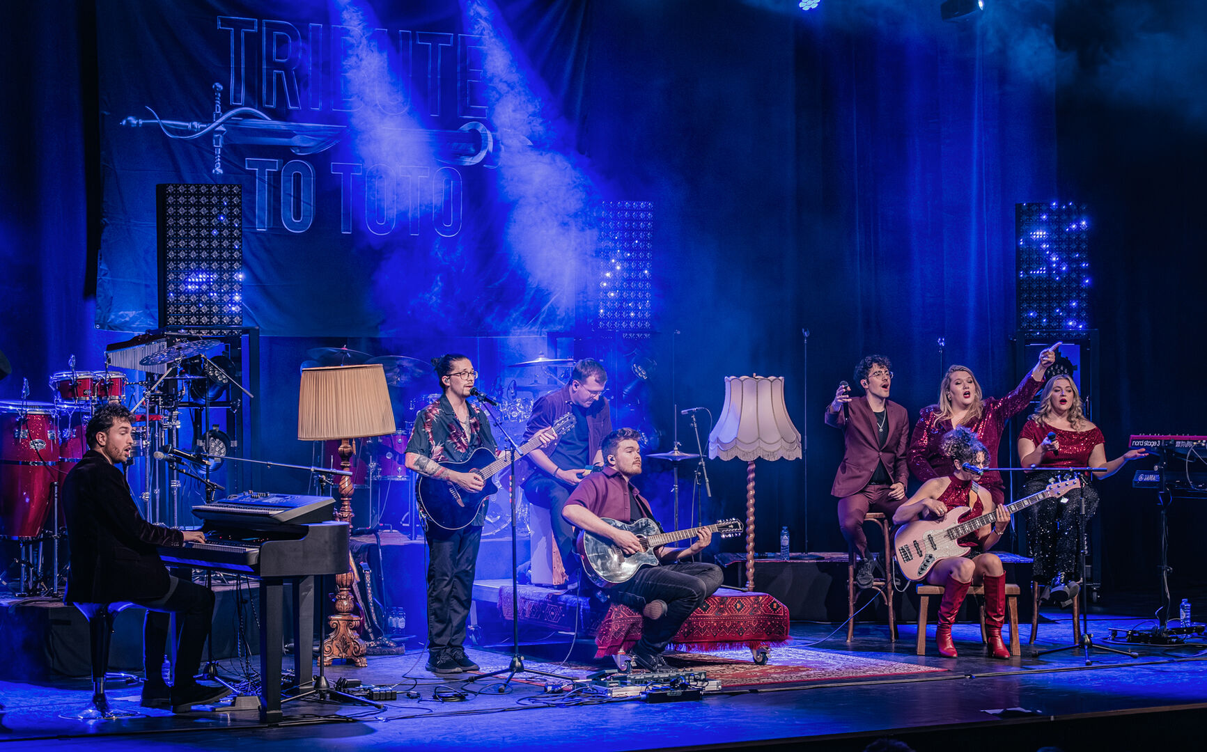 Een band speelt live op het podium, omringd door muzikanten met verschillende instrumenten en sfeervolle verlichting.