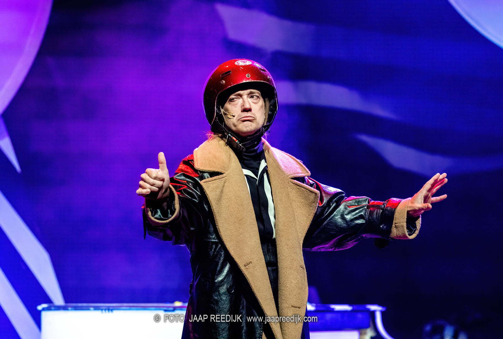 Een performer in een grote leren jas en helm staat op een podium met een kleurrijke achtergrond.
