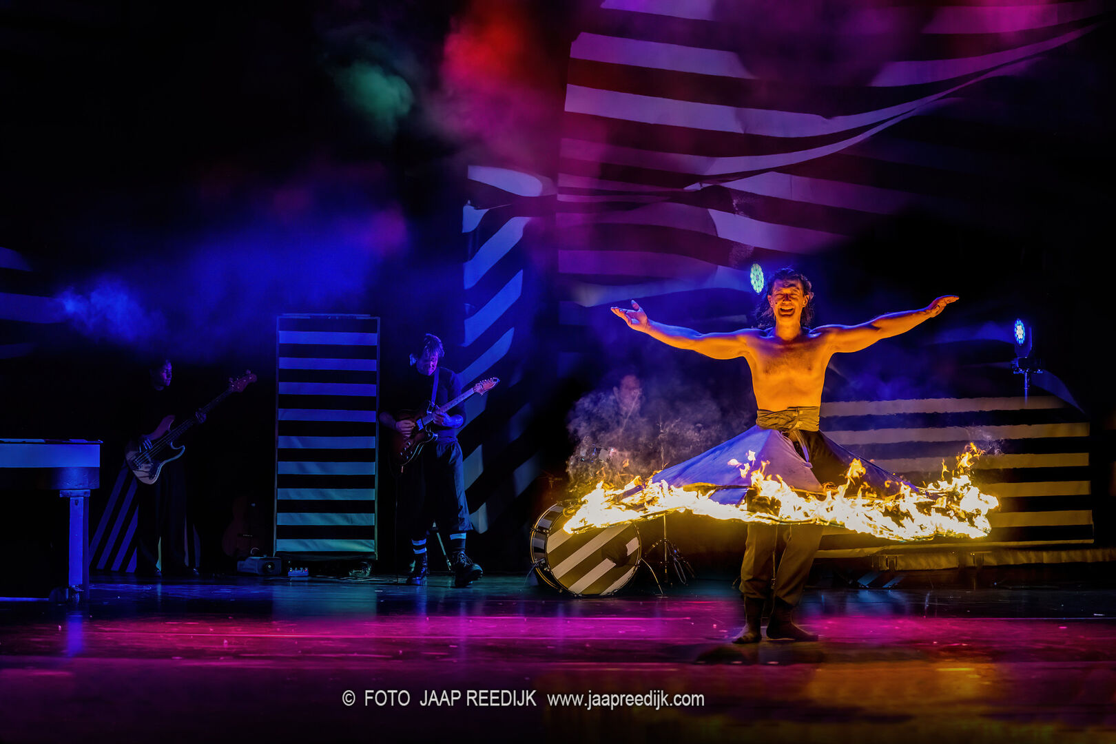 Een performer danst met een vuurcirkel op het podium, omringd door kleurrijke verlichting en een band op de achtergrond.