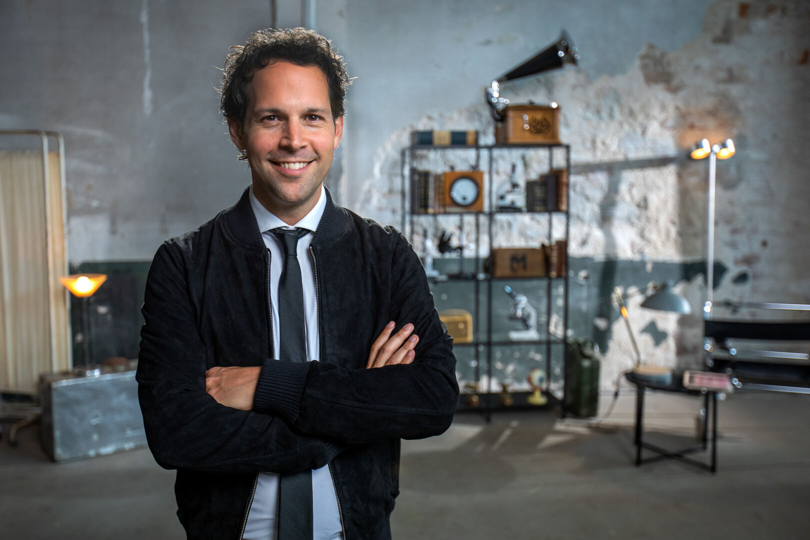 Man met een glimlach en gekruiste armen, staande in een creatieve, moderne ruimte met decoratieve elementen op de achtergrond.