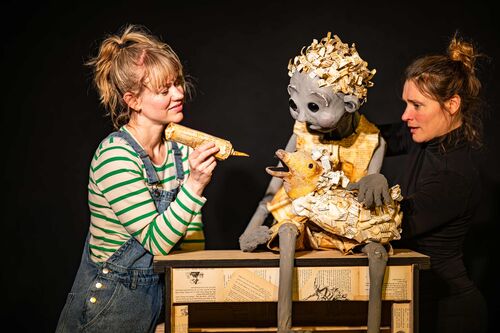 Twee vrouwen, gekleed in casual kleding, begeleiden een marionet met een gouden outfit op een houten tafel.
