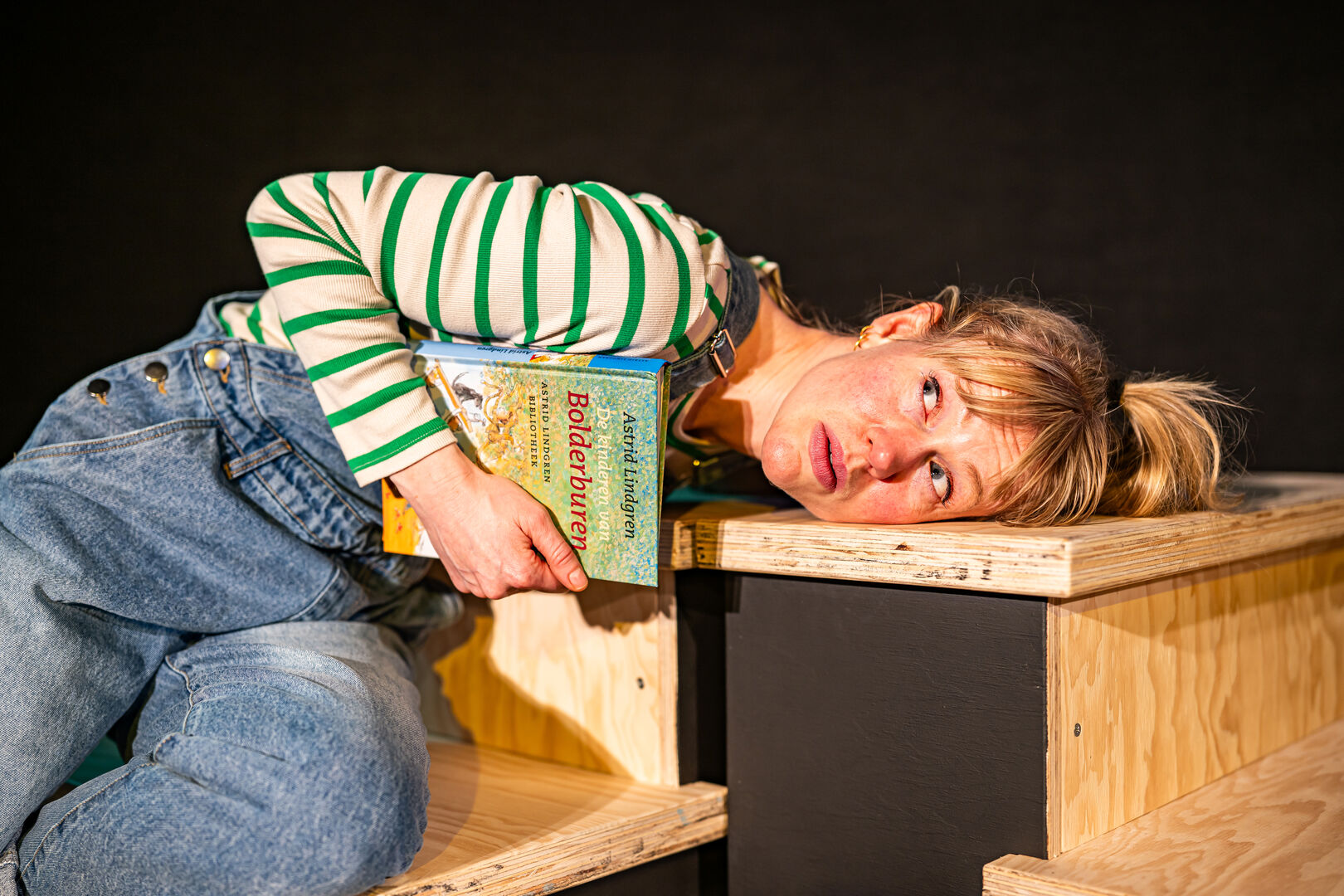Vrouw in gestreepte trui leunt op een houten plank en houdt een boek vast, met een vermoeide uitdrukking.