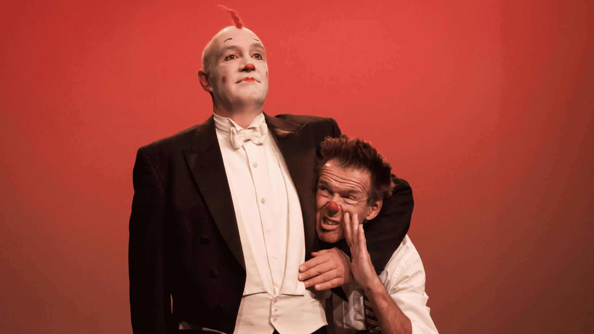 Twee acteurs op een podium; de ene in avondkleding met een witte schmink en de andere in een opvallende pose.