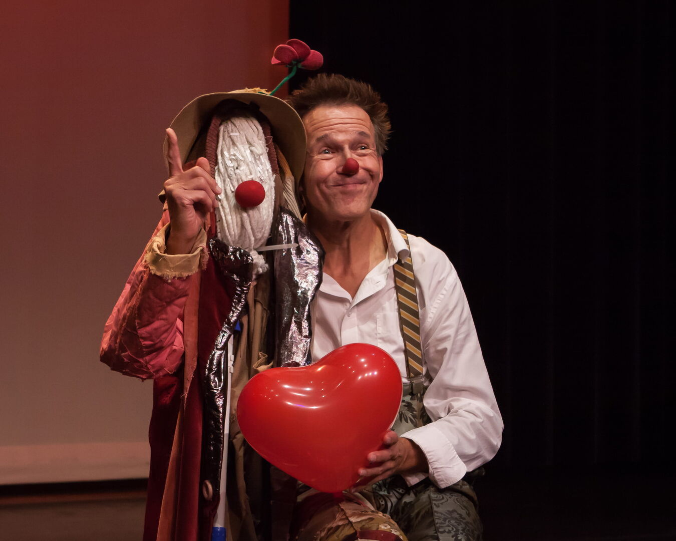 Clown met een grote rode hartballon naast een man in kostuum, beiden stralend en lachend op een podium.