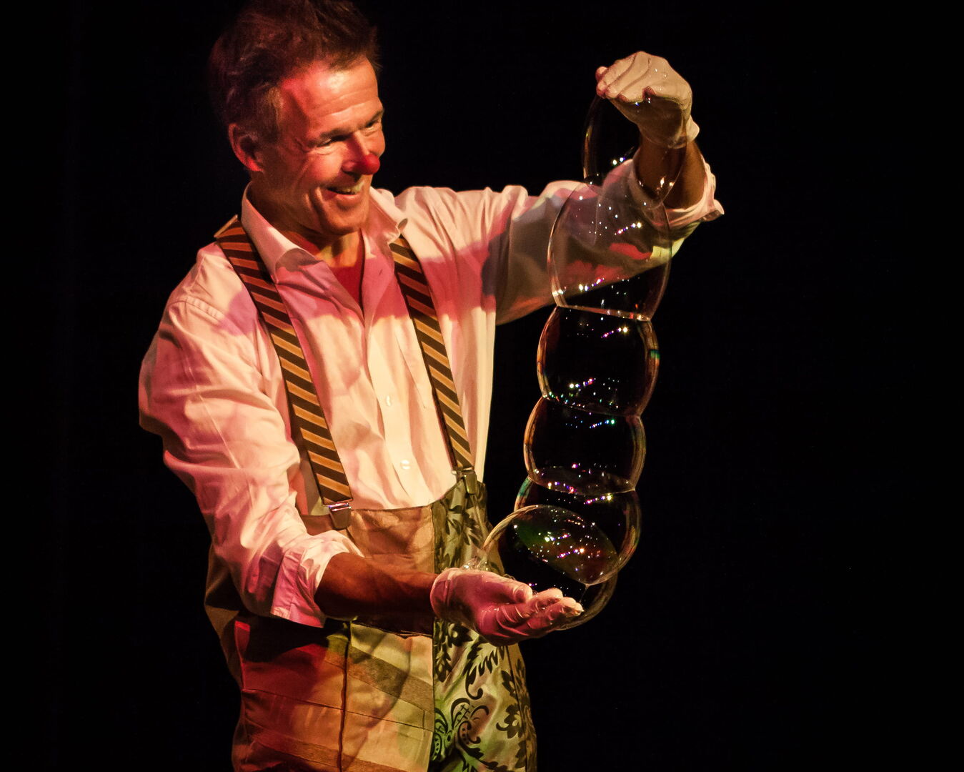 Een clown houdt meerdere zeepbellen vast terwijl hij op een podium staat, met een kleurrijke verlichting achter hem.