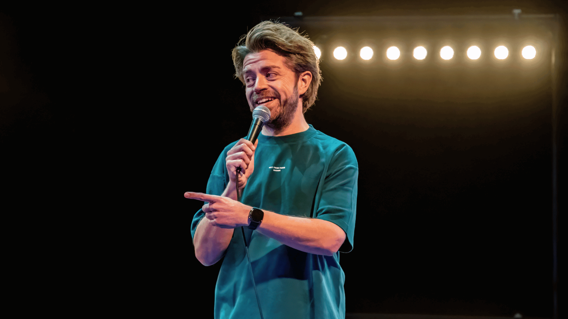 Daan van der Hoeven op het podium met microfoon in zijn hand, hij kijkt schuin weg en wijst naar het publiek
