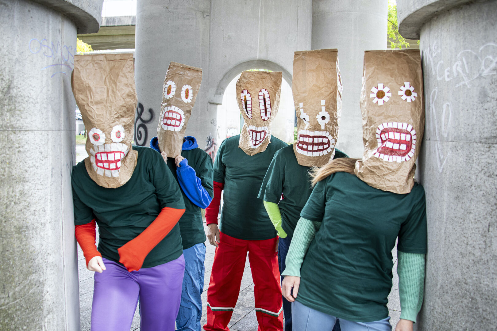 Vijf mensen met papieren zakken op hun hoofd, gekleed in groene shirts en gekleurde broeken, poseren onder een betonnen structuur.