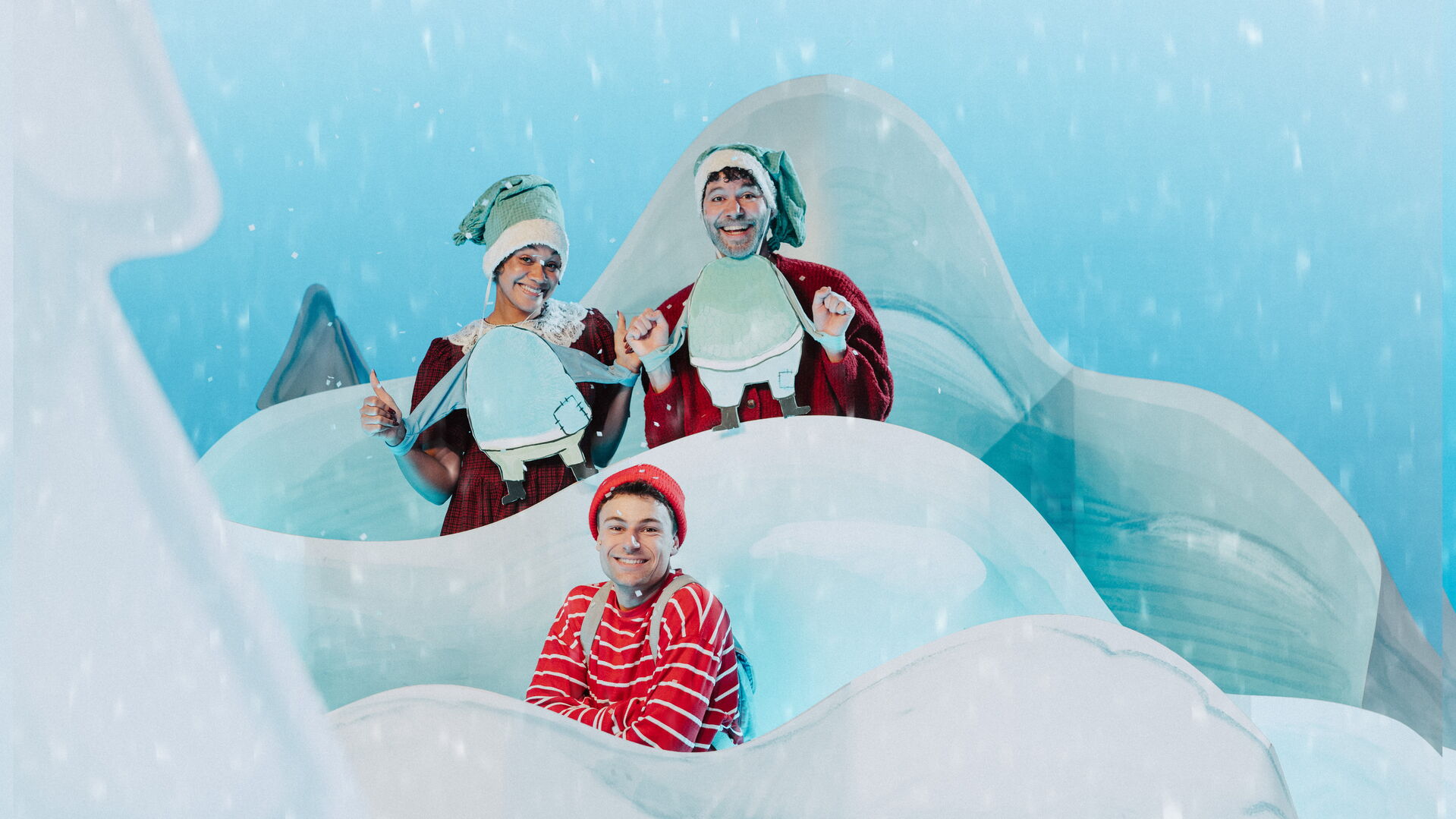 Drie feestelijk geklede mensen staan op een besneeuwd, wit decor met ijsvormen en vallen sneeuwvlokken.