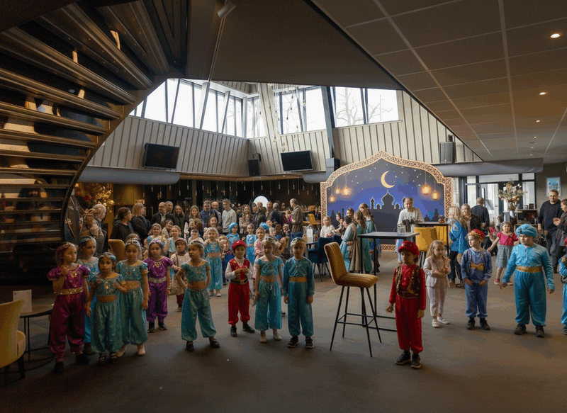 Kinderen in kleurrijke kostuums staan in een zaal, terwijl een publiek achter hen kijkt naar een voorstelling.