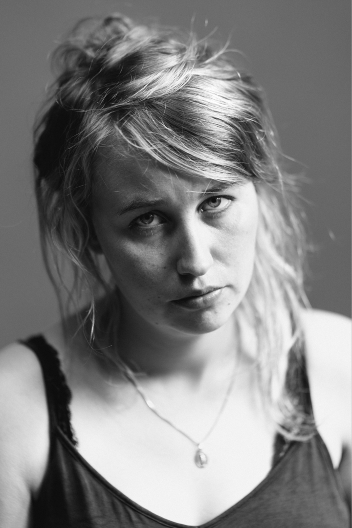 Portret van een vrouw met een serieuze uitdrukking en lang, licht gekruld haar, gefotografeerd in zwart-wit.