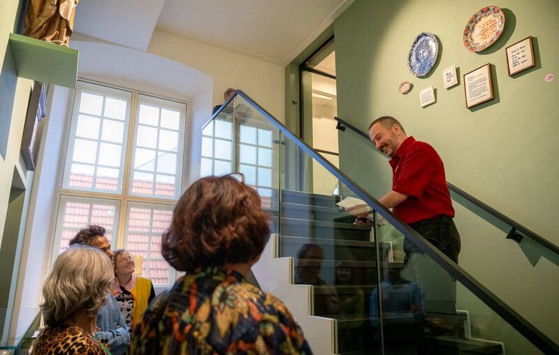 Een man in een rode trui spreekt naar een groep mensen op een trap in een lichte, moderne ruimte.