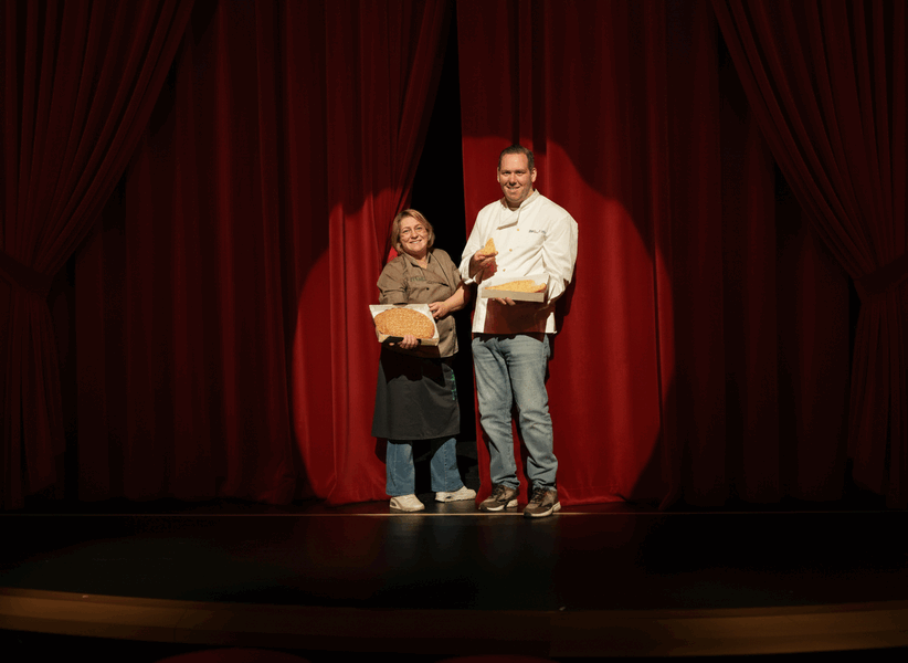 Twee personen staan op een podium met rode gordijnen, elk holding een brood, met een lichte glimlach.