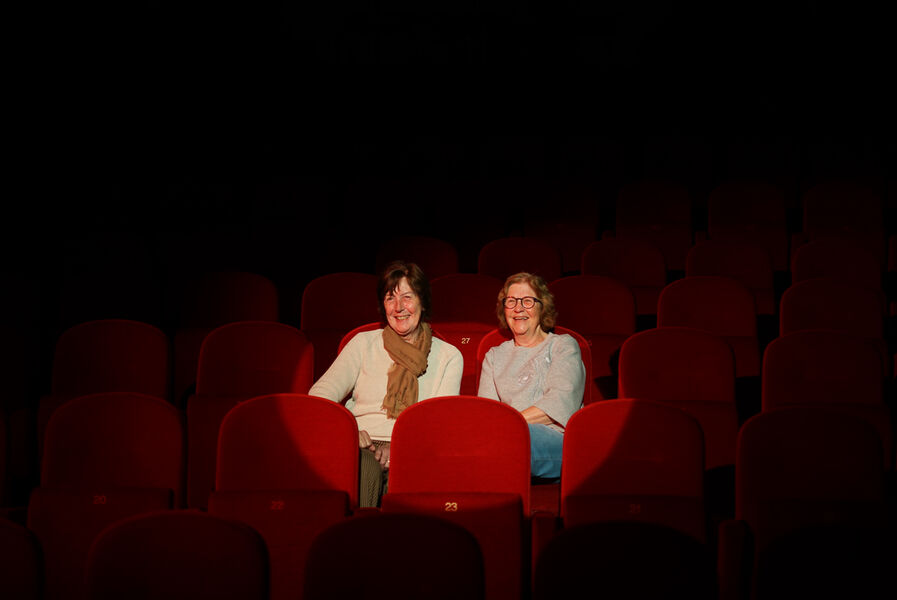 Twee vrouwen zitten samen in een lege bioscoop met rode stoelen, omringd door duistere ruimte.