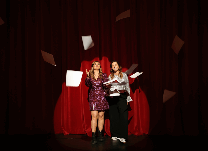Twee vrouwen in feestelijke kleding staan op een podium en laten papieren bladen in de lucht vallen tegen een rode achtergrond.