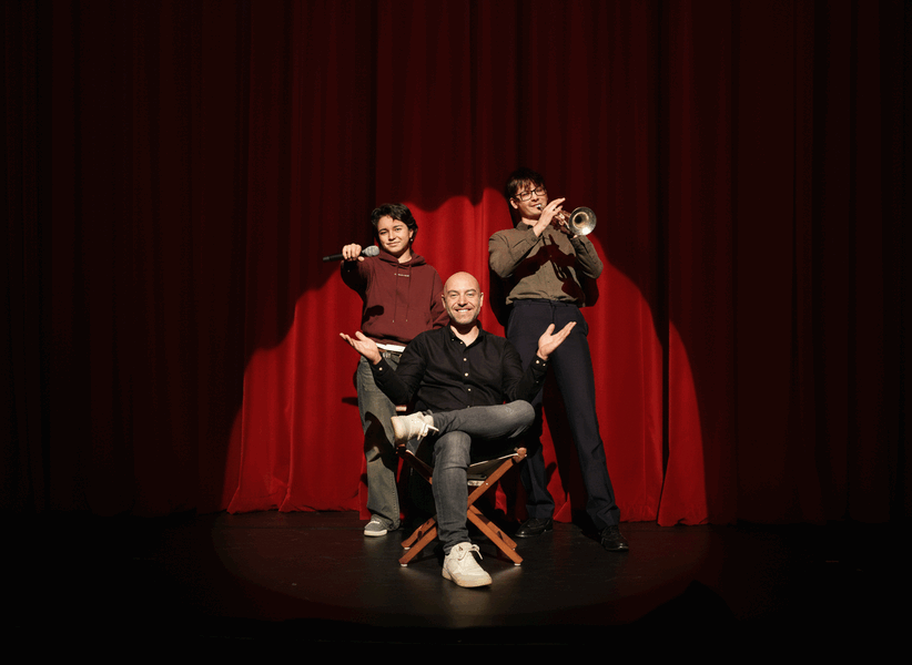 Een man in het midden op een stoel met twee spelers aan weerszijden, voor een rode theatergordijn.