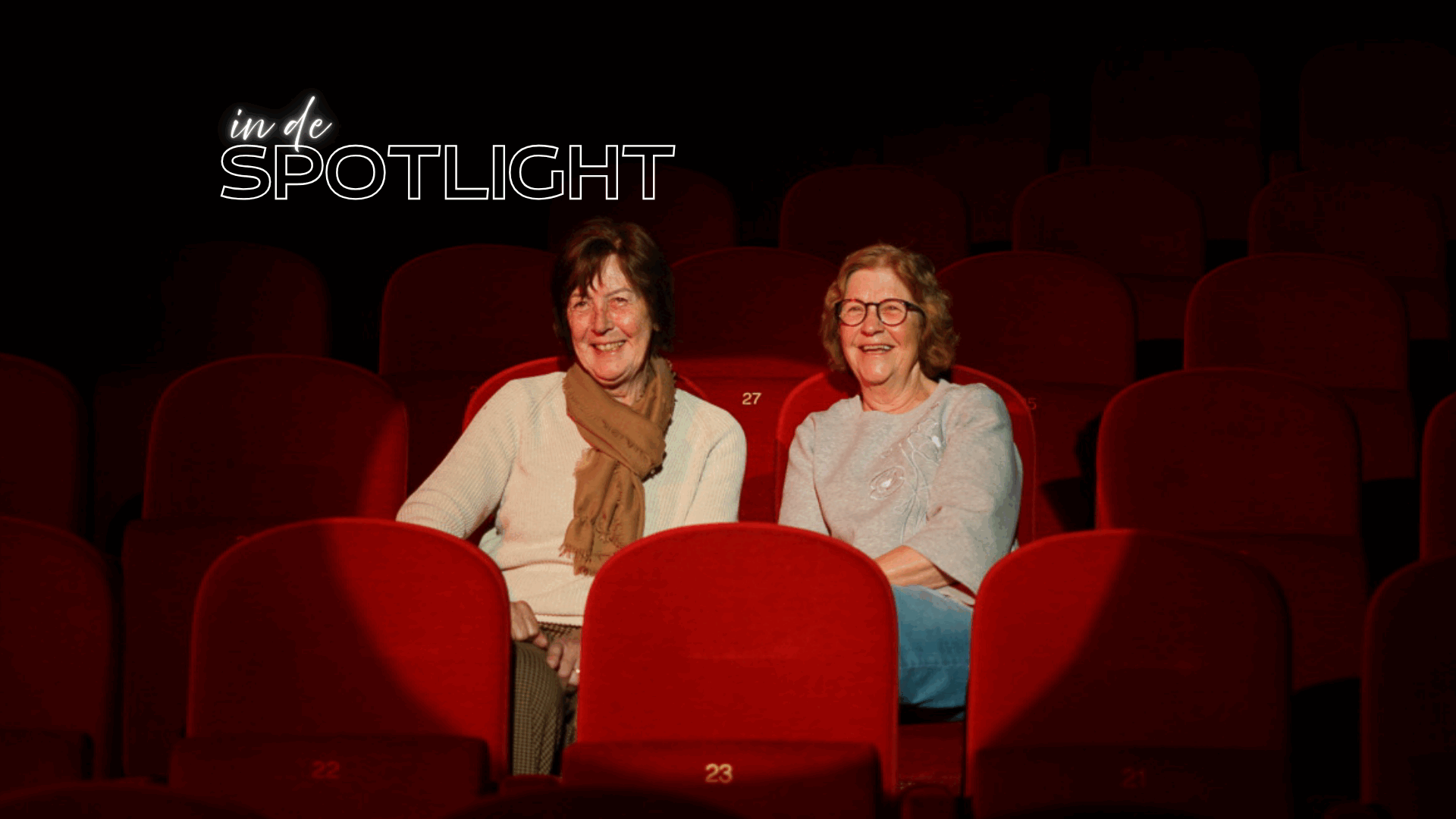 Twee vrouwen zitten op een rode theaterstoel, met een spotlight op hen gericht, te midden van lege stoelen.