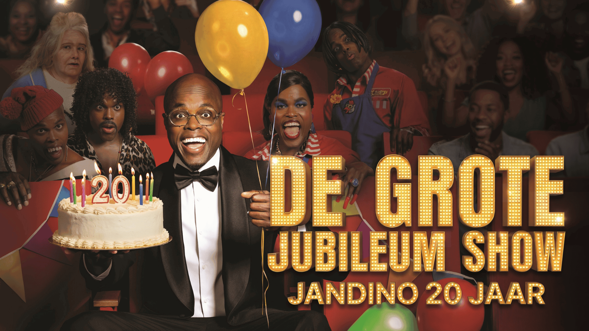 Jandino met ballonnen en een taart in zijn hand, zittend in een theaterzaal met allemaal andere spelers om hem heen