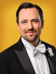 Een man in een tuxedo met een strik en bloem, glimlachend op een gele achtergrond.