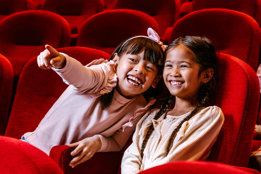 Twee lachende meisjes zitten in een theater, omringd door rode stoelen, terwijl één van hen naar iets wijst.