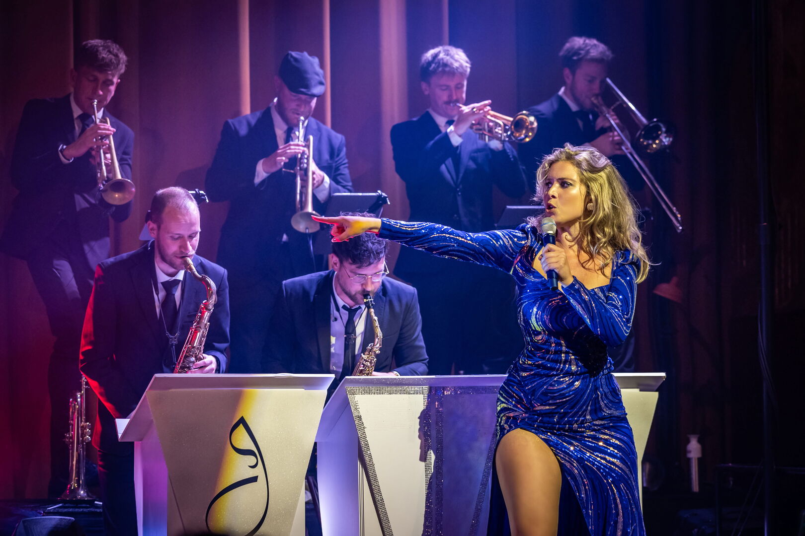 Zangeres in een blauwe jurk zingt op het podium, omringd door een band met muzikanten die blaasinstrumenten spelen.