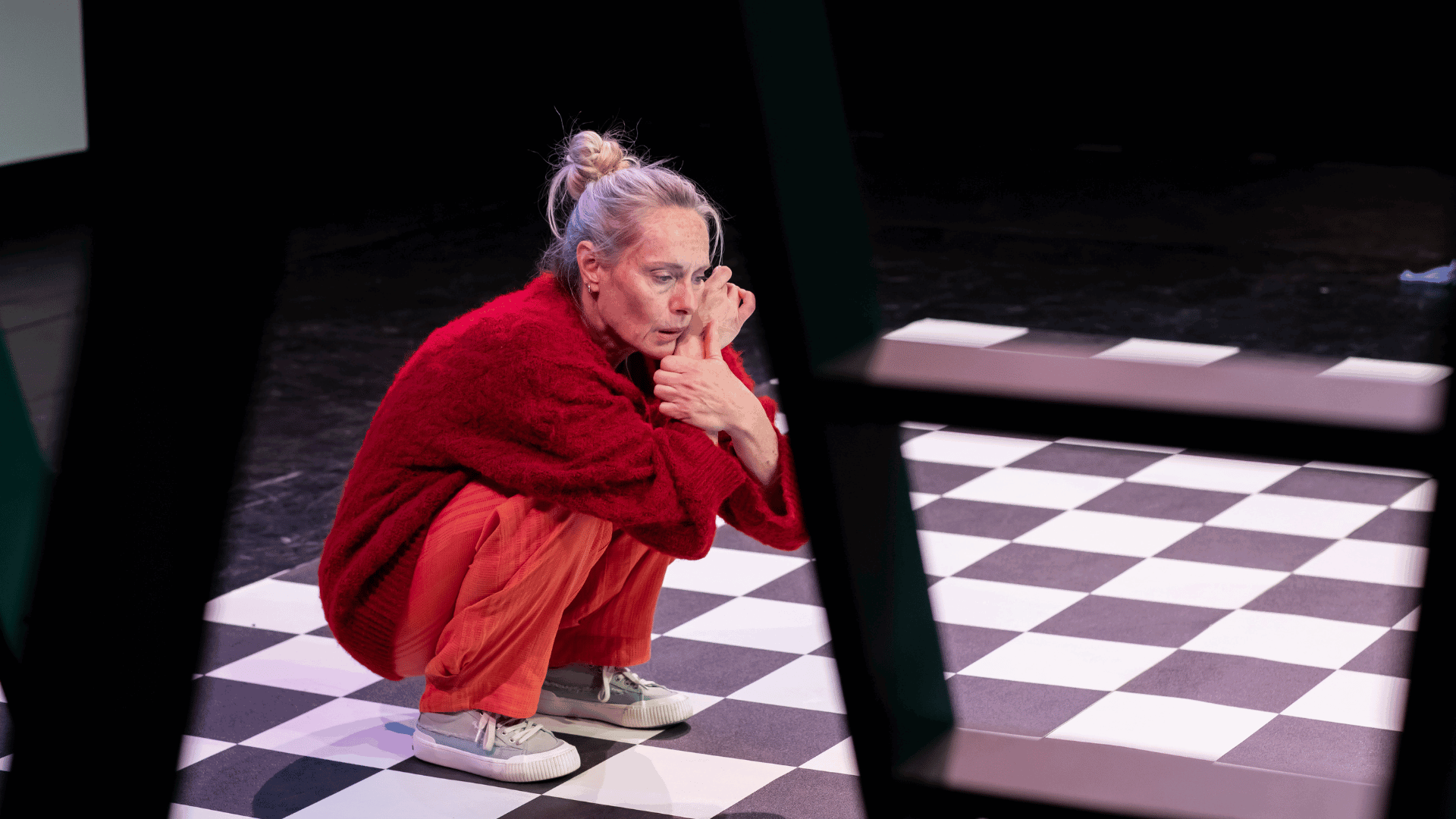 Een vrouw in een rode trui en oranje broek zit op de vloer van een theater met een zwart-wit geblokte ondergrond.