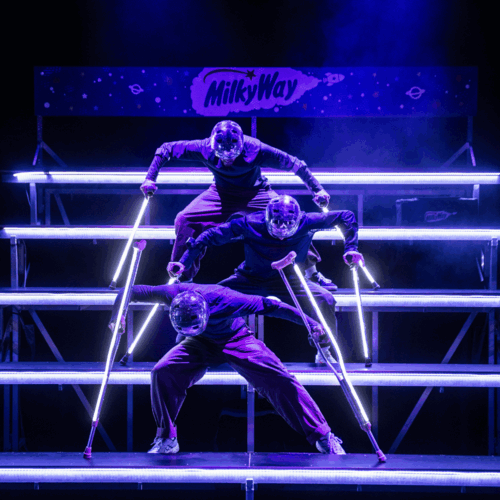 Drie acrobaten in paarse outfits performen op een verlichte trap met LED-lampen, gebruikmakend van krukken.