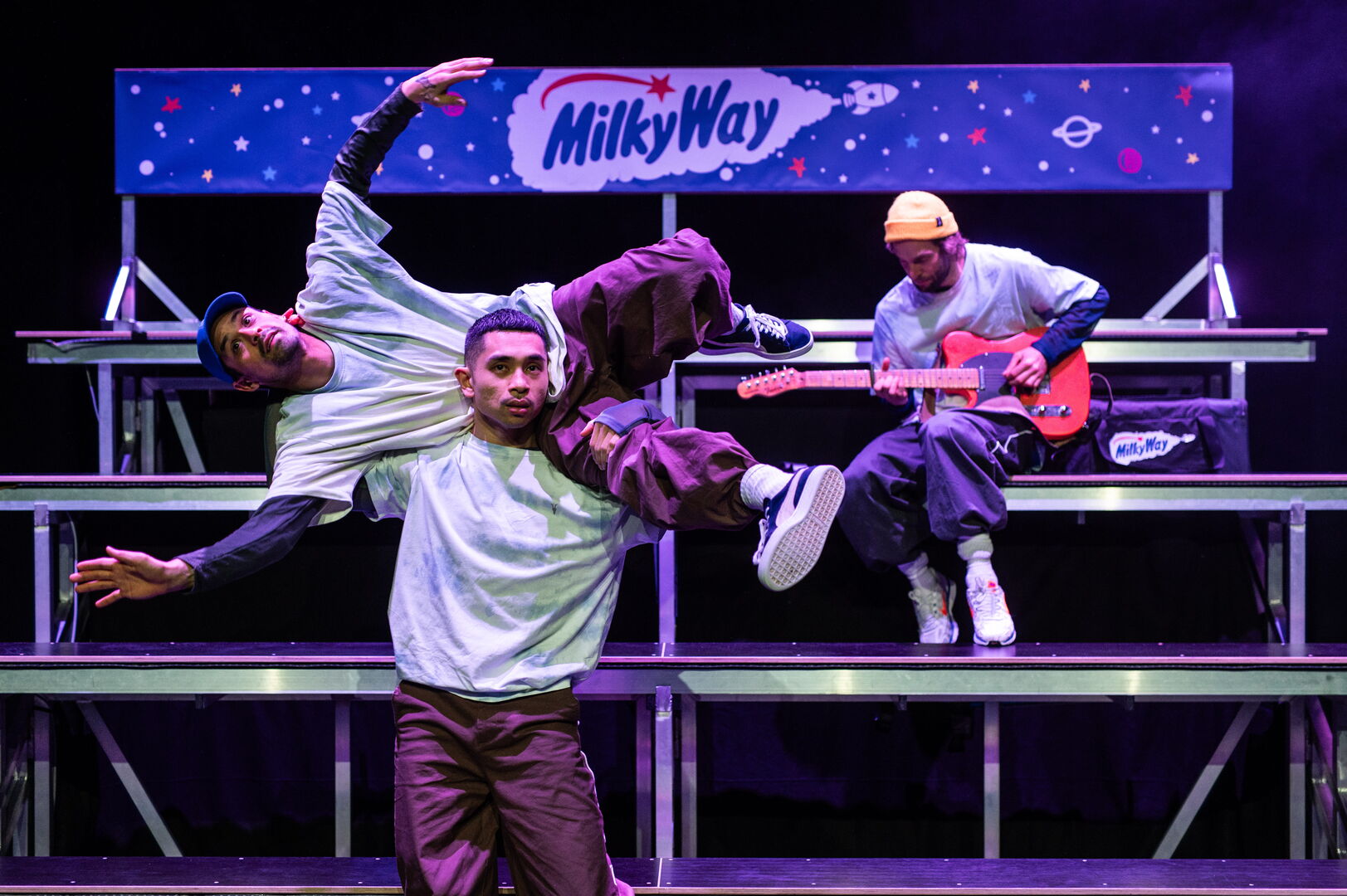Drie dansers tijdens een optreden, met een muzikant op de achtergrond die gitaar speelt.