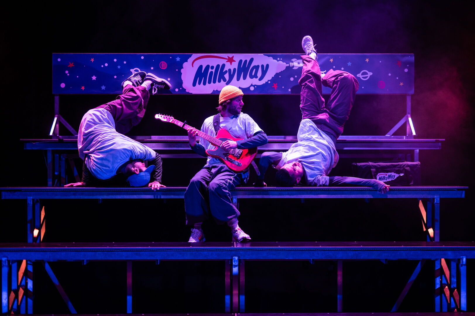 Drie performers op een verhoogd podium, één met een gitaar, gekleed in speelse kostuums, met een kleurrijke achtergrond.