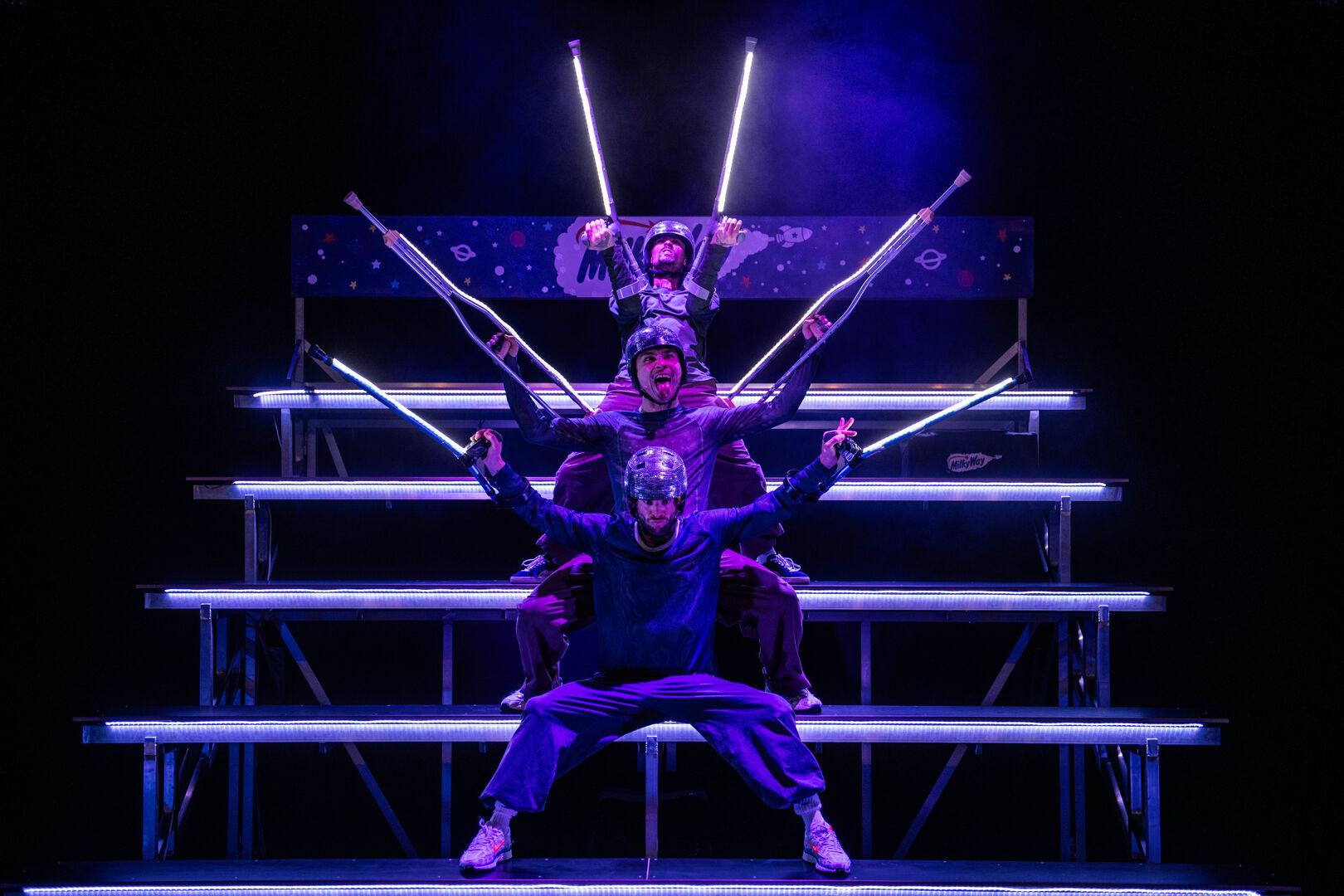 Drie dansers in paarse kostuums voeren een energieke act op met lichten en stokken op een trap.