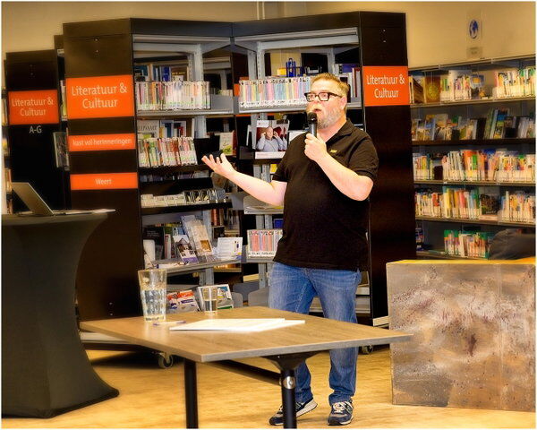 Man met een microfoon spreekt voor een publiek in een bibliotheek, met boekenplanken op de achtergrond.