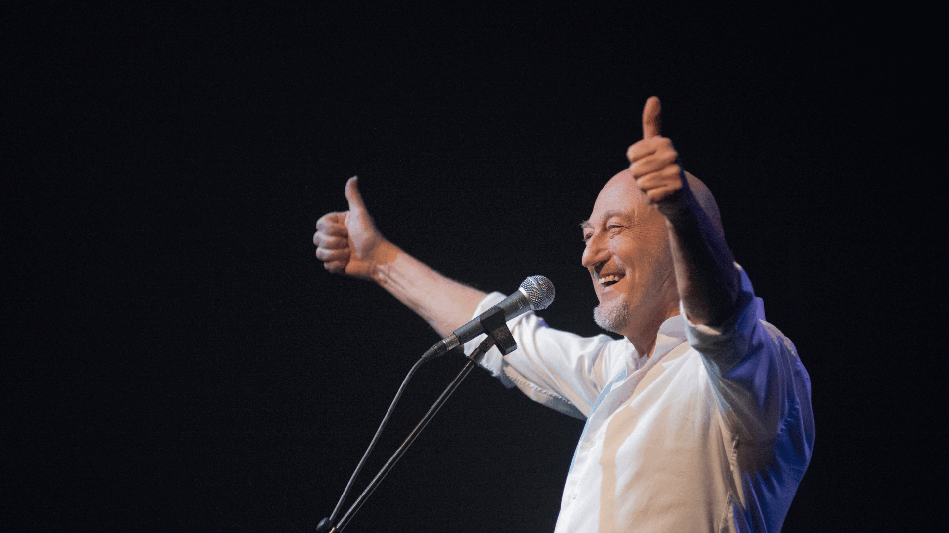 Gerard van Maasakker staat lachend achter zijn microfoon en steekt twee duimen op