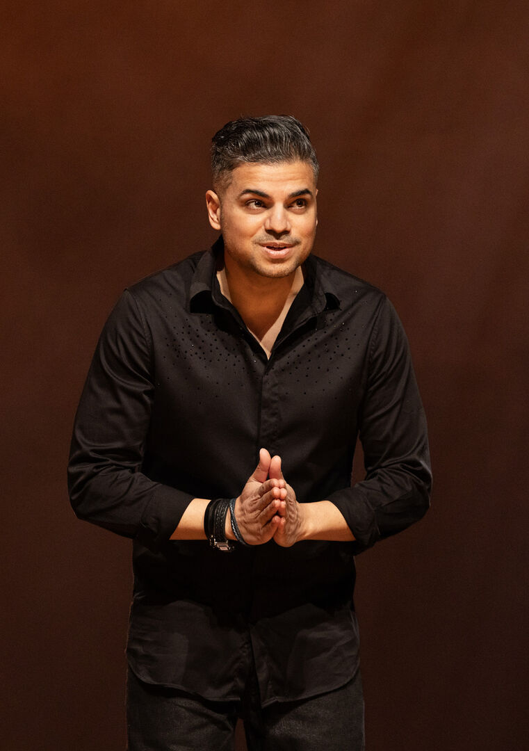 Comedian op een podium, gekleed in zwart, met een expressieve houding en handen samengevoegd in een gebedsgeste.