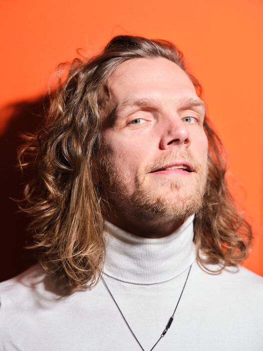 Portret van een man met lang, golvend haar, gekleed in een hoge kraag, tegen een oranje achtergrond.