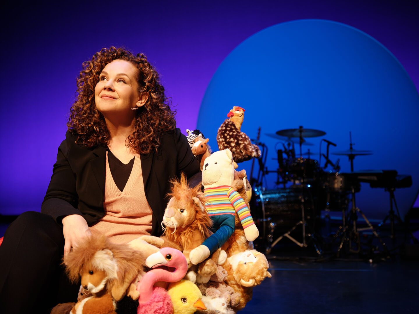 Vrouw met krullend haar zit op het podium omringd door knuffeldieren en kijkt dromerig naar de achtergrond.