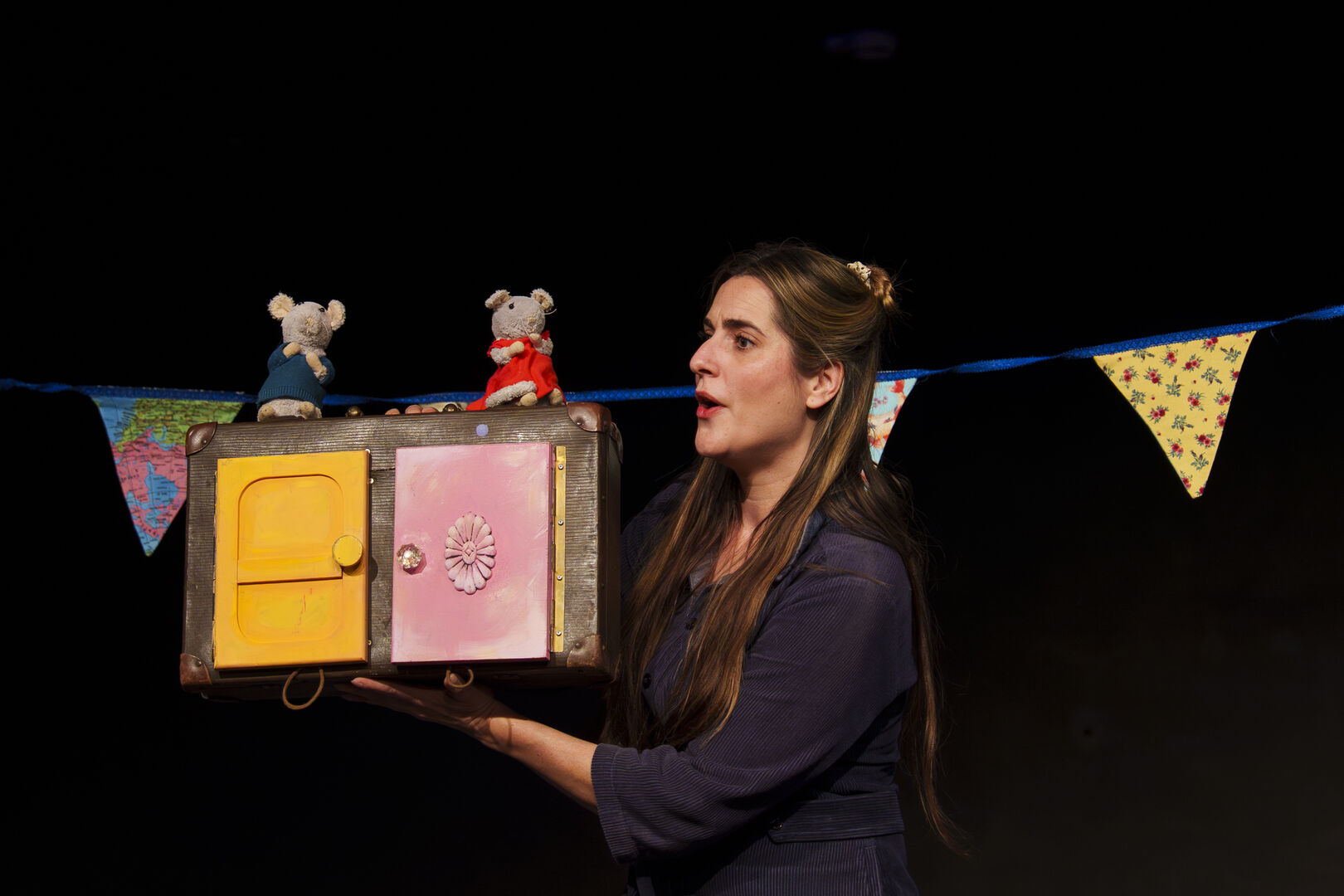 Een vrouw toont een kleurrijk speelgoedkastje met twee muisfiguren bovenop, terwijl ze vertelt op een podium.