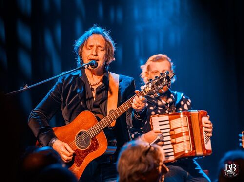 Twee muzikanten optreden op een podium; een bespeelt een gitaar en de ander een accordeon. Verlichte achtergrond.