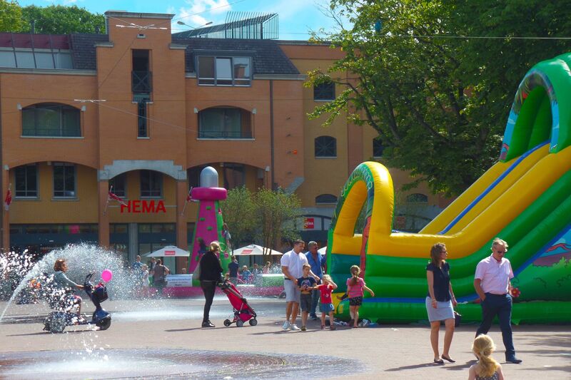 Op een zomerse dag spelen kinderen bij een luchtkussen en waterfonteinen op een plein. Achtergrond met gebouwen en mensen.
