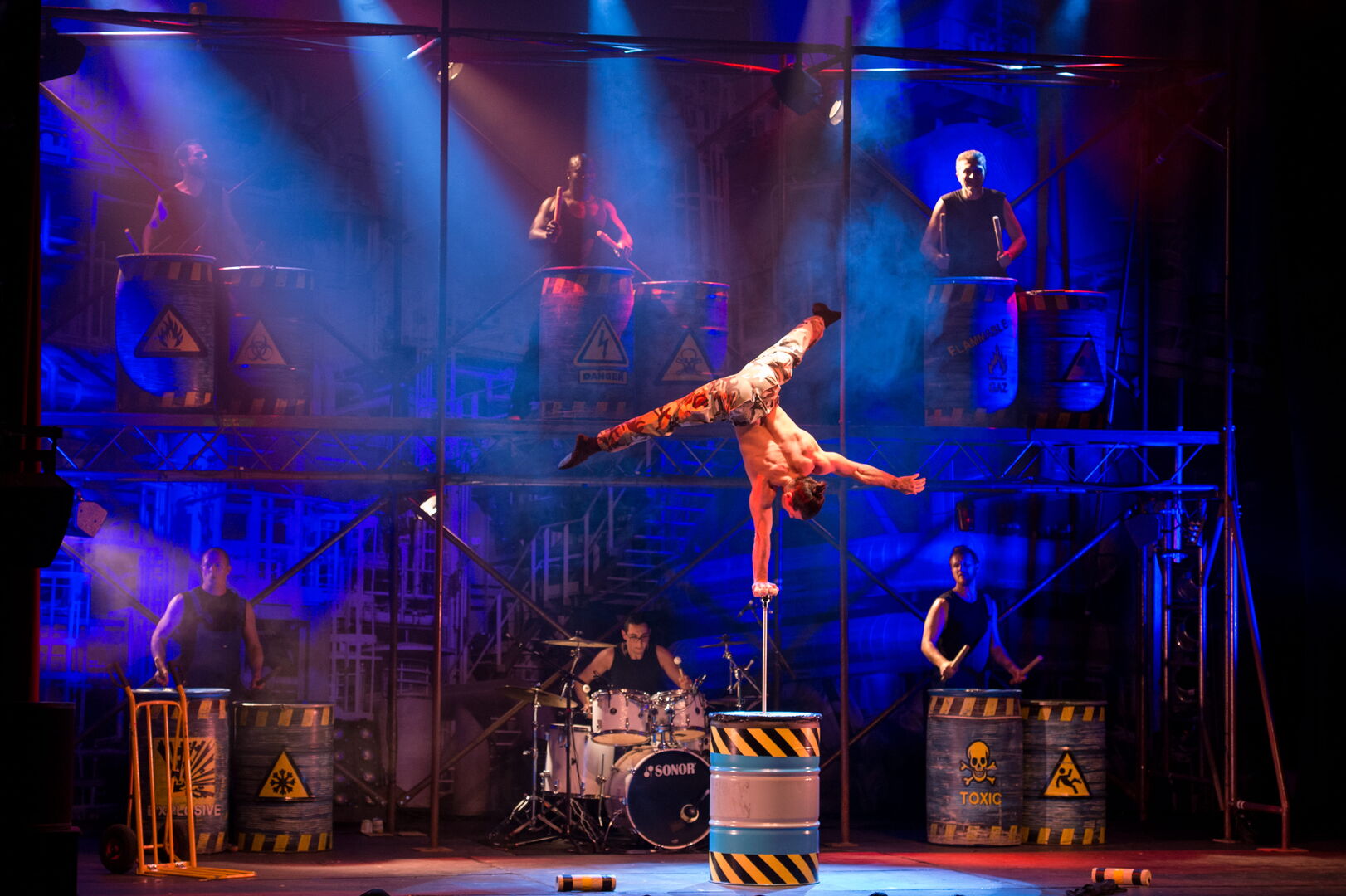 Een performer voert een acrobatische act uit op een metalen barrel, omringd door muzikanten op het podium.