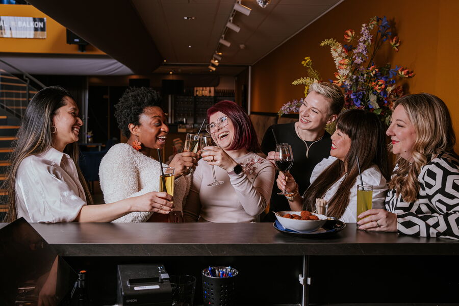 Groep vrouwen in een bar proostend met drankjes, omringd door een kleurrijke inrichting en bloemen.