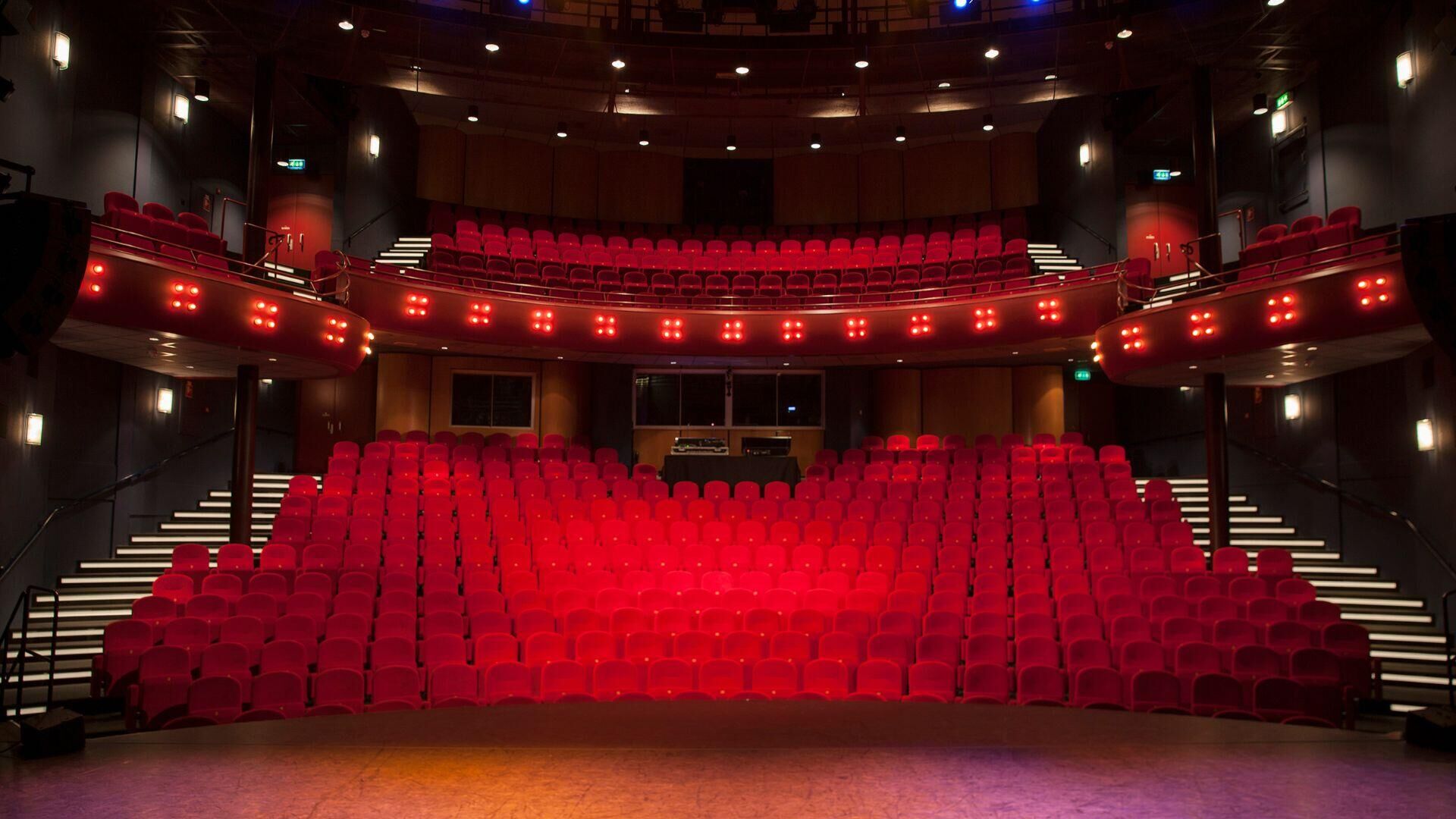 In een theater met rode stoelen en een podium, verlicht door enkele lichten, klaar voor een voorstelling.