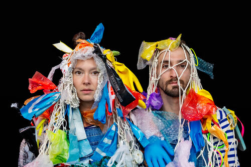 Twee personen met kleding en accessoires gemaakt van plastic afval, tegen een zwarte achtergrond.